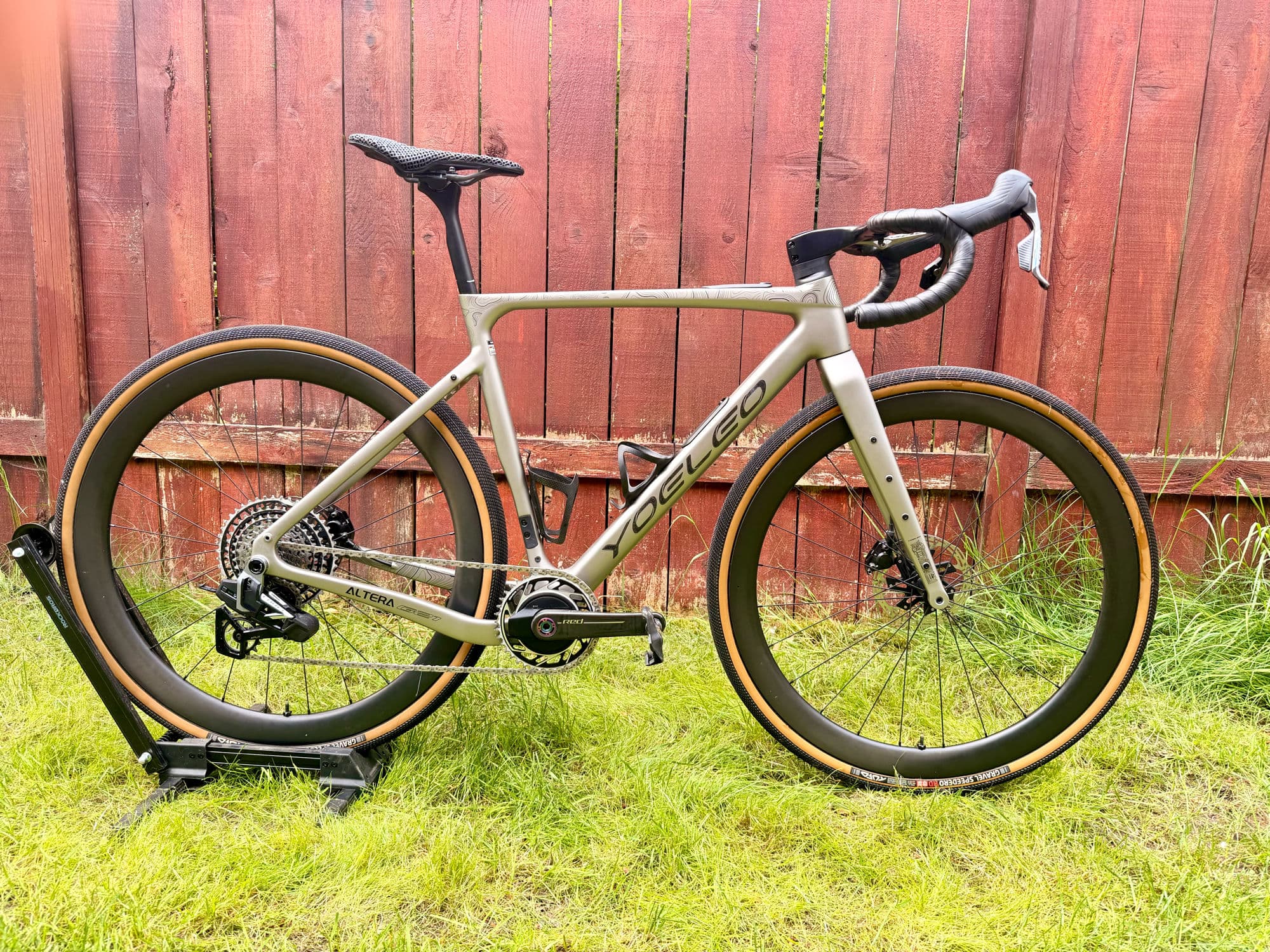 A champagne Yoeleo Altera G21 gravel frame on a workshop stand against a red cedar fence — SAT C45 carbon wheels with tan-wall Speedero tires and SRAM Force XPLR E1 with a Quarq spider power meter.
